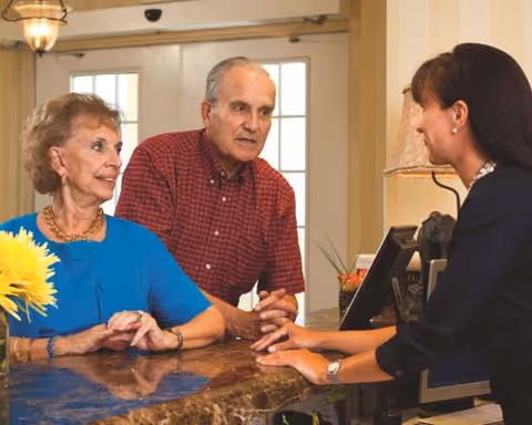 An elderly couple talking to a receptionist at a front desk inside a facility. The woman is wearing a blue top and the man is wearing a red checkered shirt. The receptionist is smiling and engaging with them. There is a vase with yellow flowers on the counter and a lamp in the background.