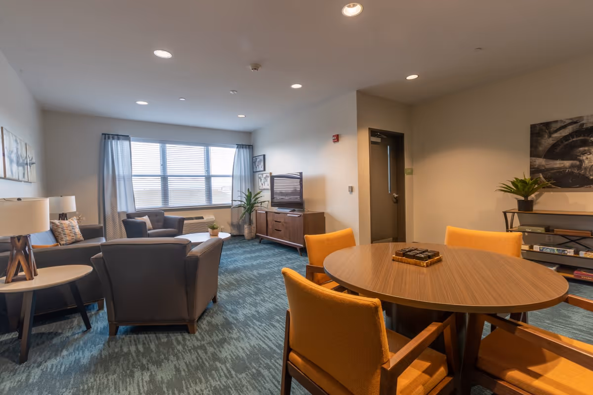 Communal living room with a seating area, television, and a round table surrounded by orange chairs.