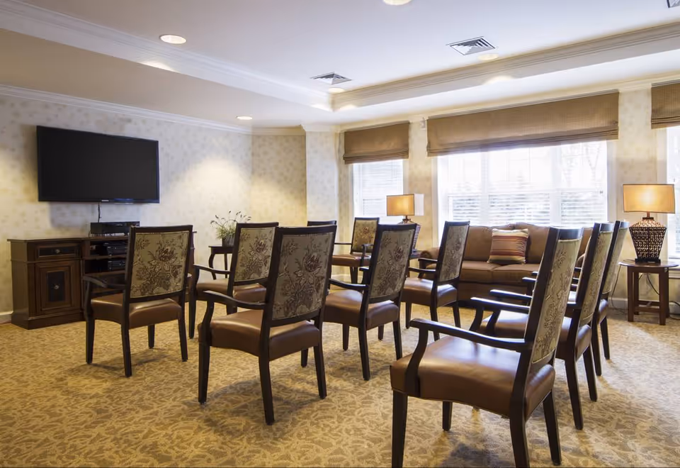 A well-lit common room with multiple wooden chairs arranged in rows facing a wall-mounted flat screen TV. The room has beige patterned carpet, light-colored wallpaper, large windows with blinds, a brown sofa with a striped pillow, and two side tables with lamps.