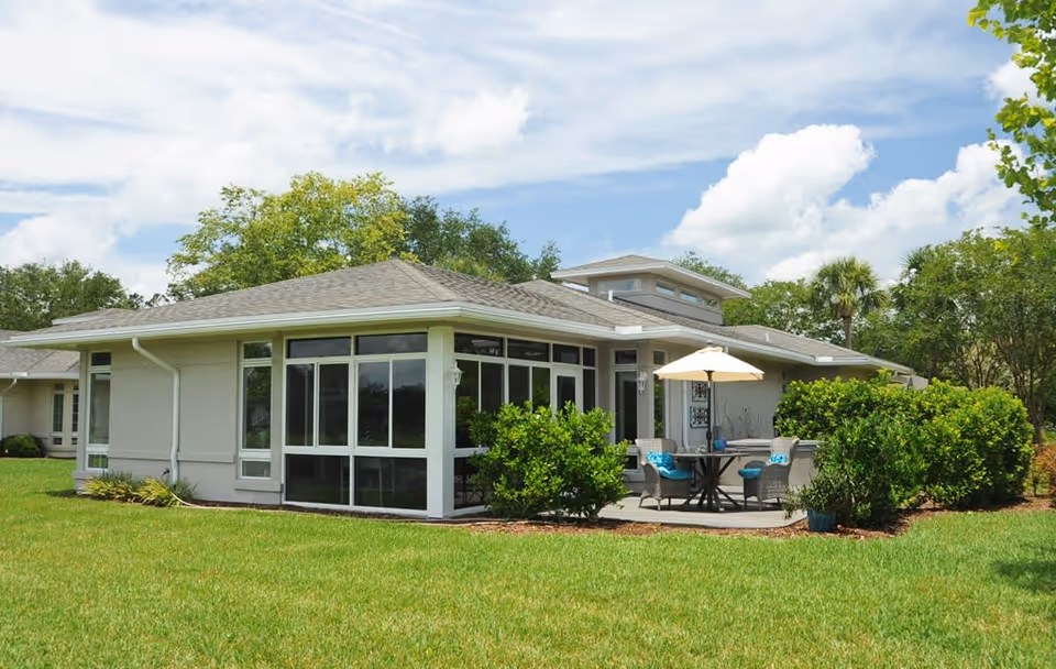 Exterior view of a single-story building with large windows, surrounded by green grass and bushes under a partly cloudy sky. There is a patio area with a table, chairs, and an umbrella.