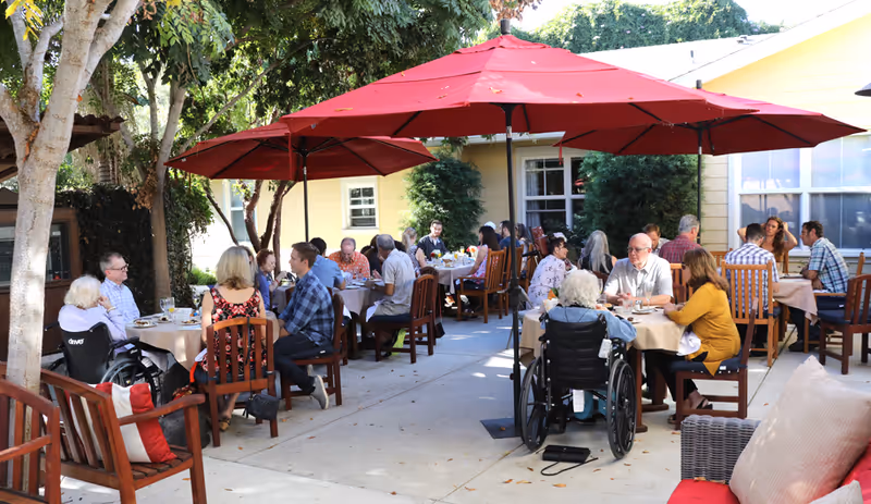 Outdoor patio area at Heritage House-An Assisted Living Community with multiple tables where elderly residents and visitors are seated, enjoying a meal under large red umbrellas. The setting is shaded by trees and surrounded by a yellow building with windows.