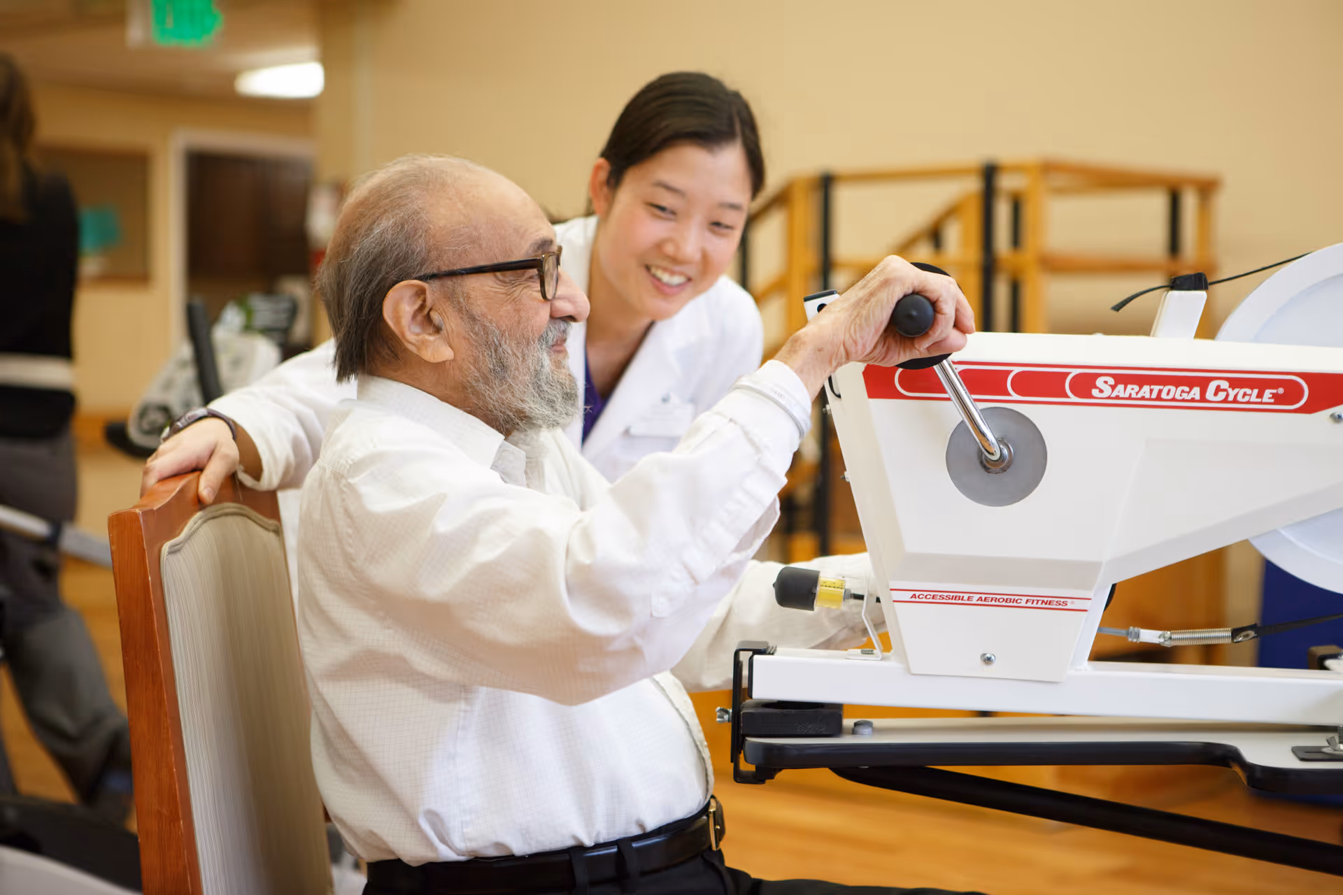 An elderly man with glasses is seated and using a Saratoga Cycle exercise machine while a smiling female caregiver or therapist in a white coat supports and encourages him in a well-lit indoor setting.