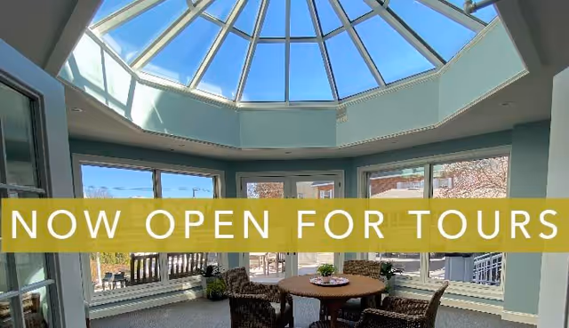 A bright indoor seating area with a round table and four wicker chairs under a large glass skylight. Large windows surround the space, showing an outdoor patio and greenery. A yellow banner across the image reads 'NOW OPEN FOR TOURS'.