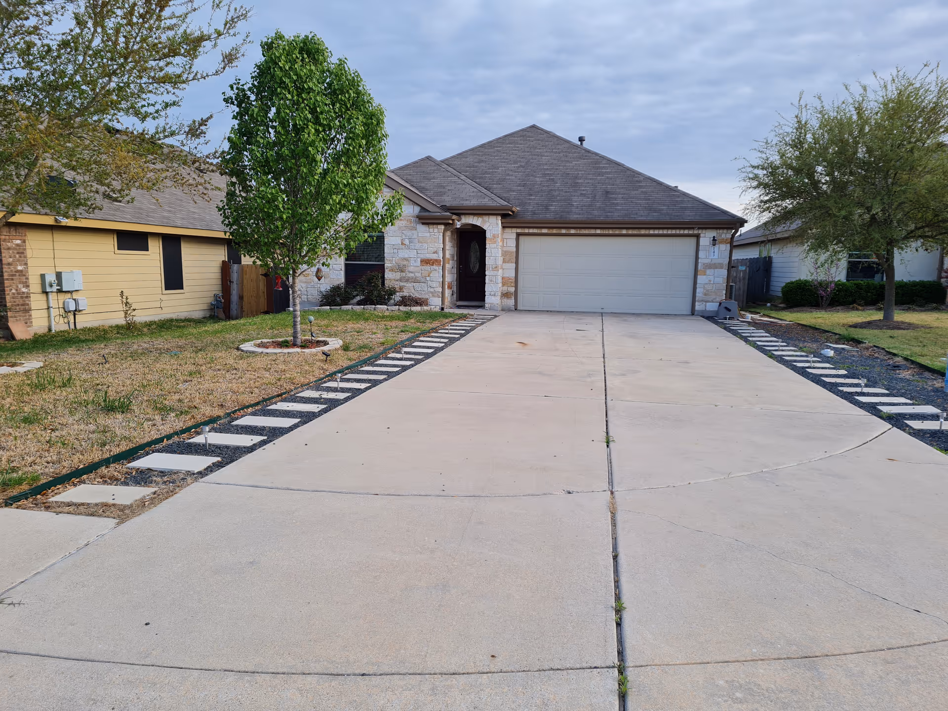 Front exterior of a single-story house with a wide concrete driveway, two-car garage, and a small front yard with trees.