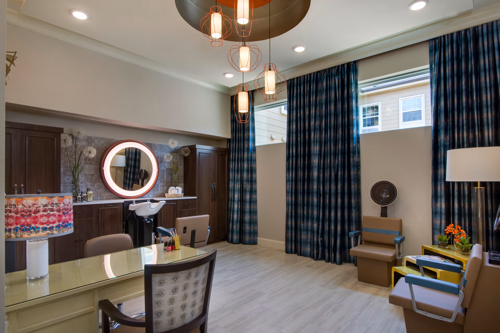 A well-lit salon room with a glass-top desk and chair in the foreground, a hair washing station with a round illuminated mirror on the back wall, dark wood cabinets, and two salon chairs with a hair dryer. Blue patterned curtains cover large windows, and modern pendant lights hang from the ceiling.