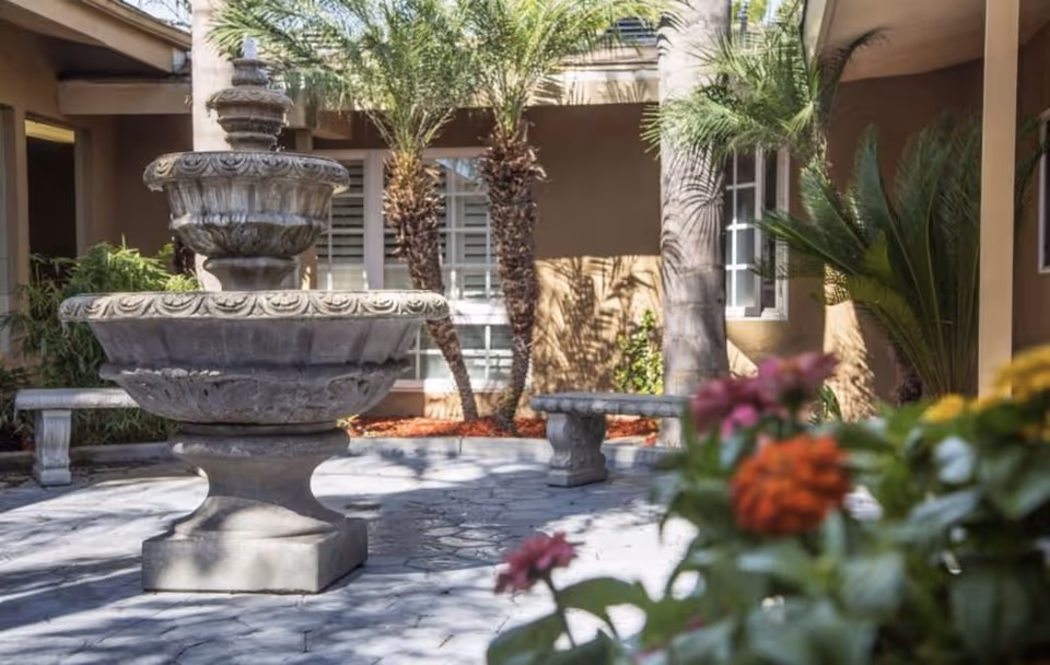 Outdoor courtyard area with a large stone fountain in the center, surrounded by palm trees and plants. There are stone benches around the fountain and a building with windows in the background. Some colorful flowers are visible in the foreground.