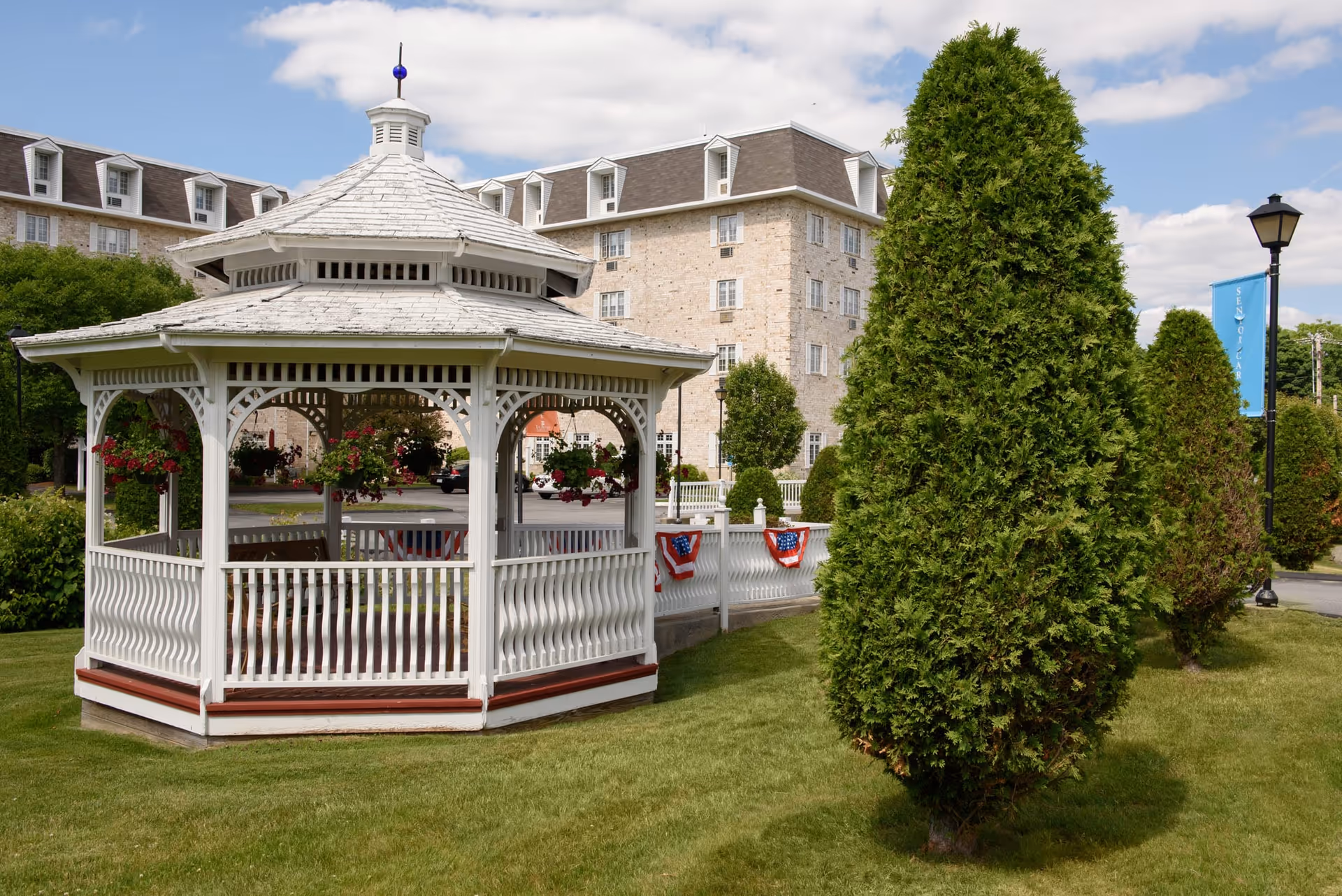 A white wooden gazebo with hanging flower baskets is situated on a well-maintained lawn with neatly trimmed bushes and trees. In the background, there is a large multi-story stone building with multiple windows under a partly cloudy sky. A white fence decorated with red, white, and blue bunting runs behind the gazebo.