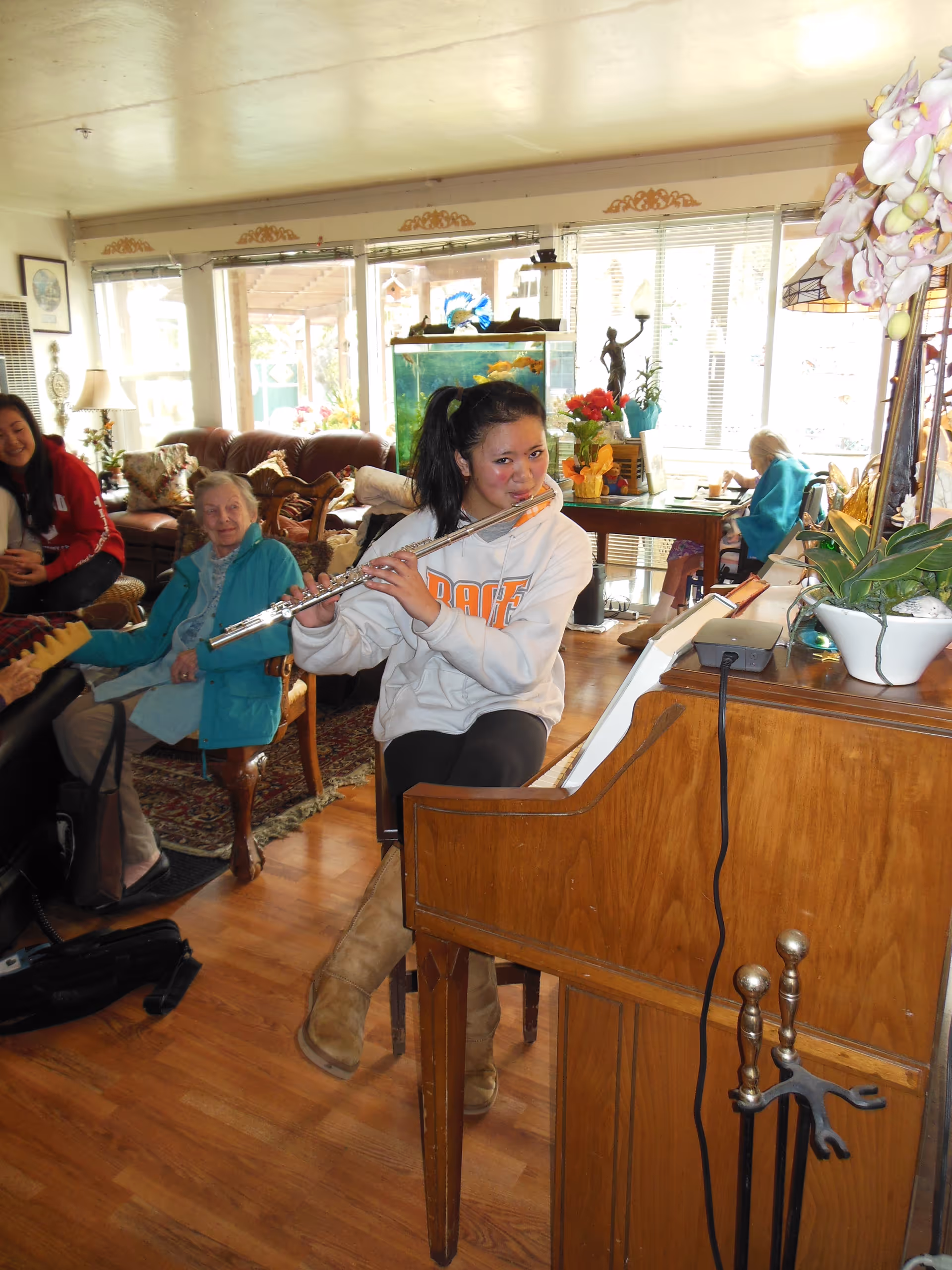 A young woman playing the flute while seated at a piano in a cozy living room. Several elderly people are seated around her, listening and smiling. The room has wooden floors, large windows with natural light, an aquarium, plants, and comfortable seating.