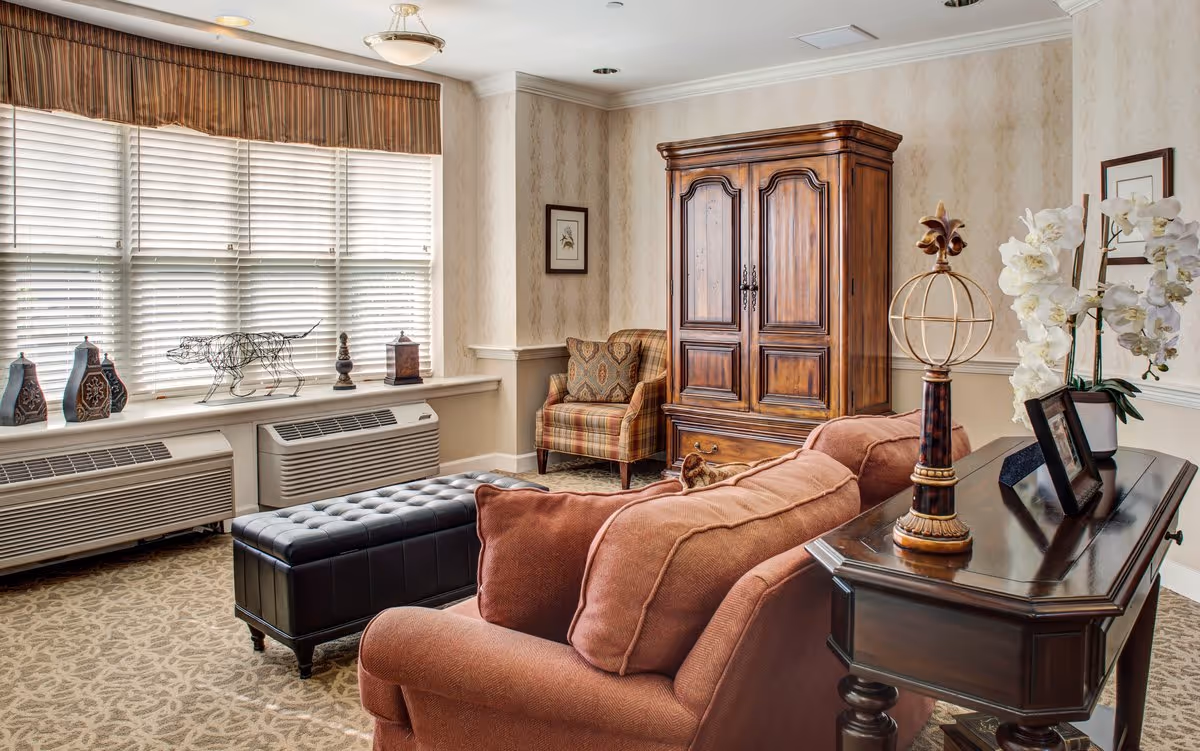 A cozy living room with a large window covered by blinds and a valance. The room features a brown upholstered sofa with cushions, a black tufted leather ottoman, a wooden armoire, and a plaid armchair with a decorative pillow. A dark wooden console table holds a decorative globe sculpture, framed photos, and a white orchid plant. The walls have a light patterned wallpaper and the floor is carpeted with a beige patterned rug.