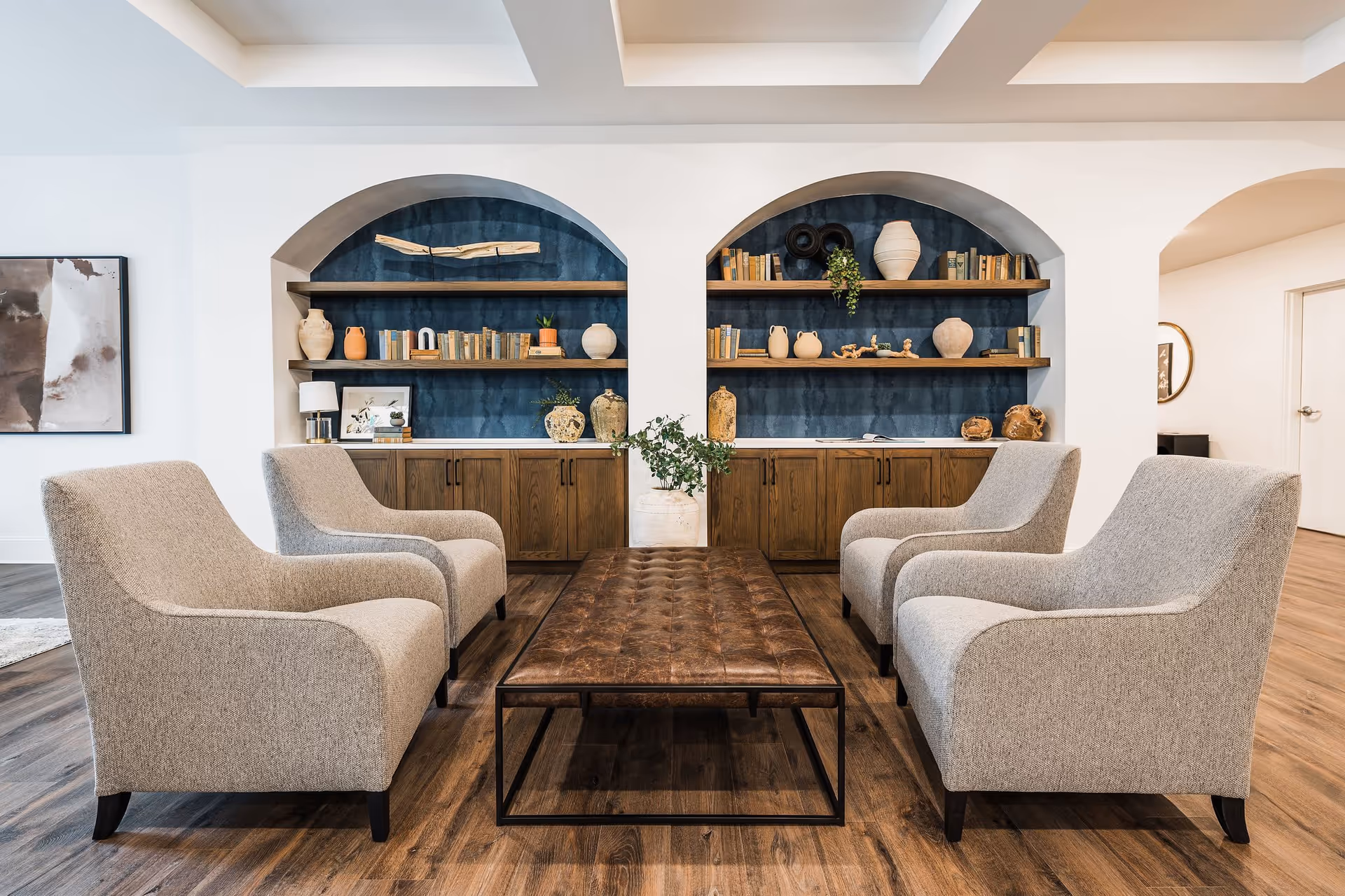 A cozy seating area with four beige upholstered armchairs arranged around a rectangular brown leather tufted coffee table. Behind the seating area are two built-in wooden shelves with decorative pottery, books, and plants against a blue textured wall. The floor is wood, and the ceiling has recessed lighting.