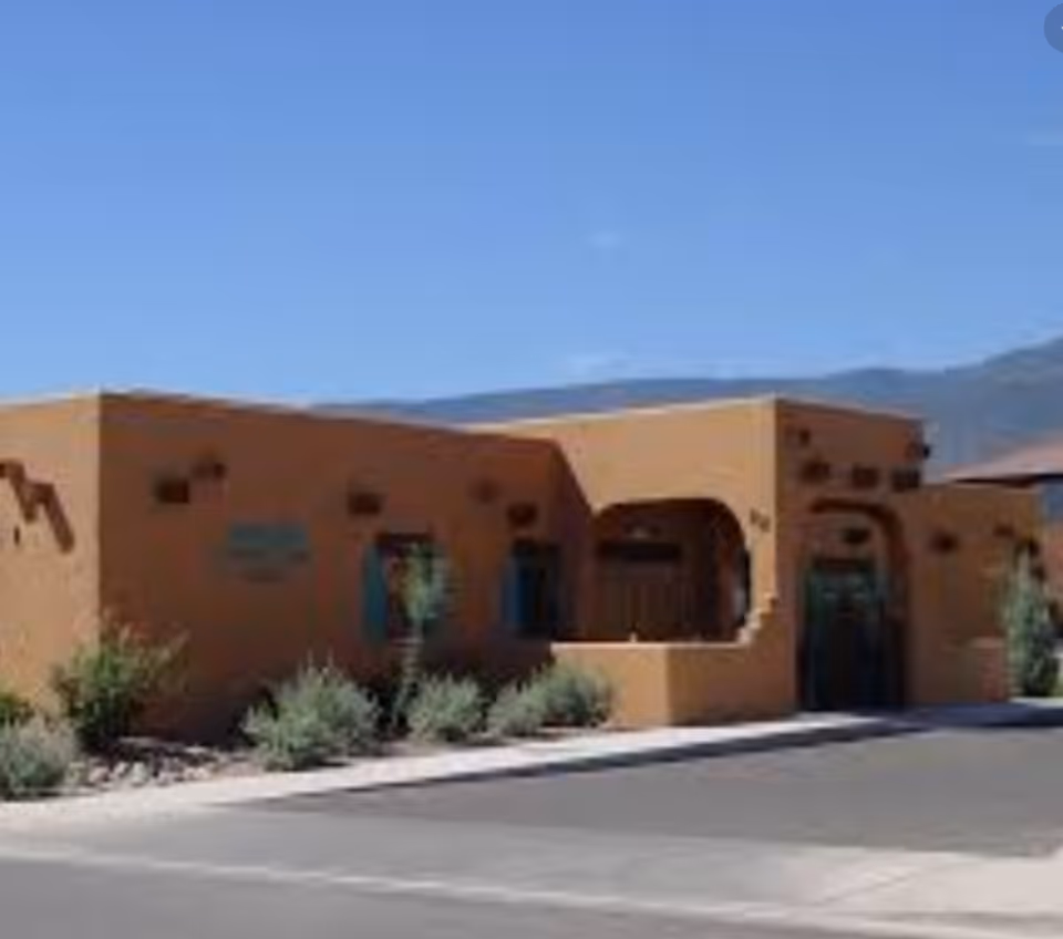 Exterior view of a single-story building with adobe-style architecture, featuring a flat roof and rounded archways. The building is surrounded by desert landscaping with shrubs and small plants, and mountains are visible in the background under a clear blue sky.