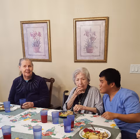 Three people sit at a dining table with plates of food and plastic cups, with two framed floral prints on the wall behind them.
