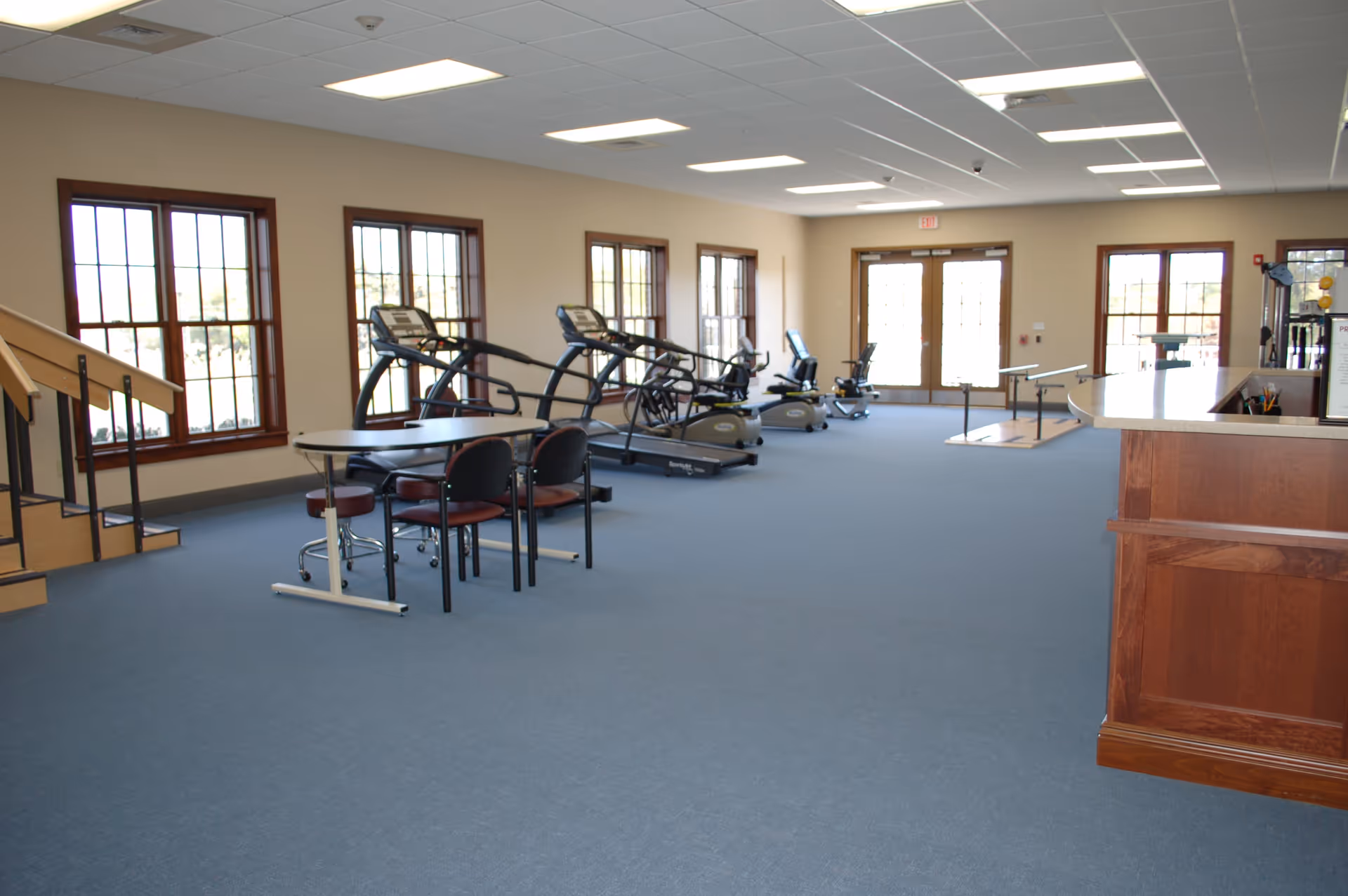 A spacious exercise room with several treadmills and stationary bikes lined up near large windows. There is a table with chairs in the foreground and a wooden reception desk on the right side. The room has blue carpet, beige walls, and fluorescent ceiling lights.