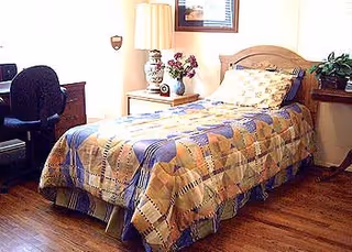A single bed with a patterned quilt in a room with wooden flooring. Next to the bed is a nightstand with a decorative lamp and a vase of flowers. There is a wooden desk with a black office chair on the left side of the image, and a window with blinds above the bed.