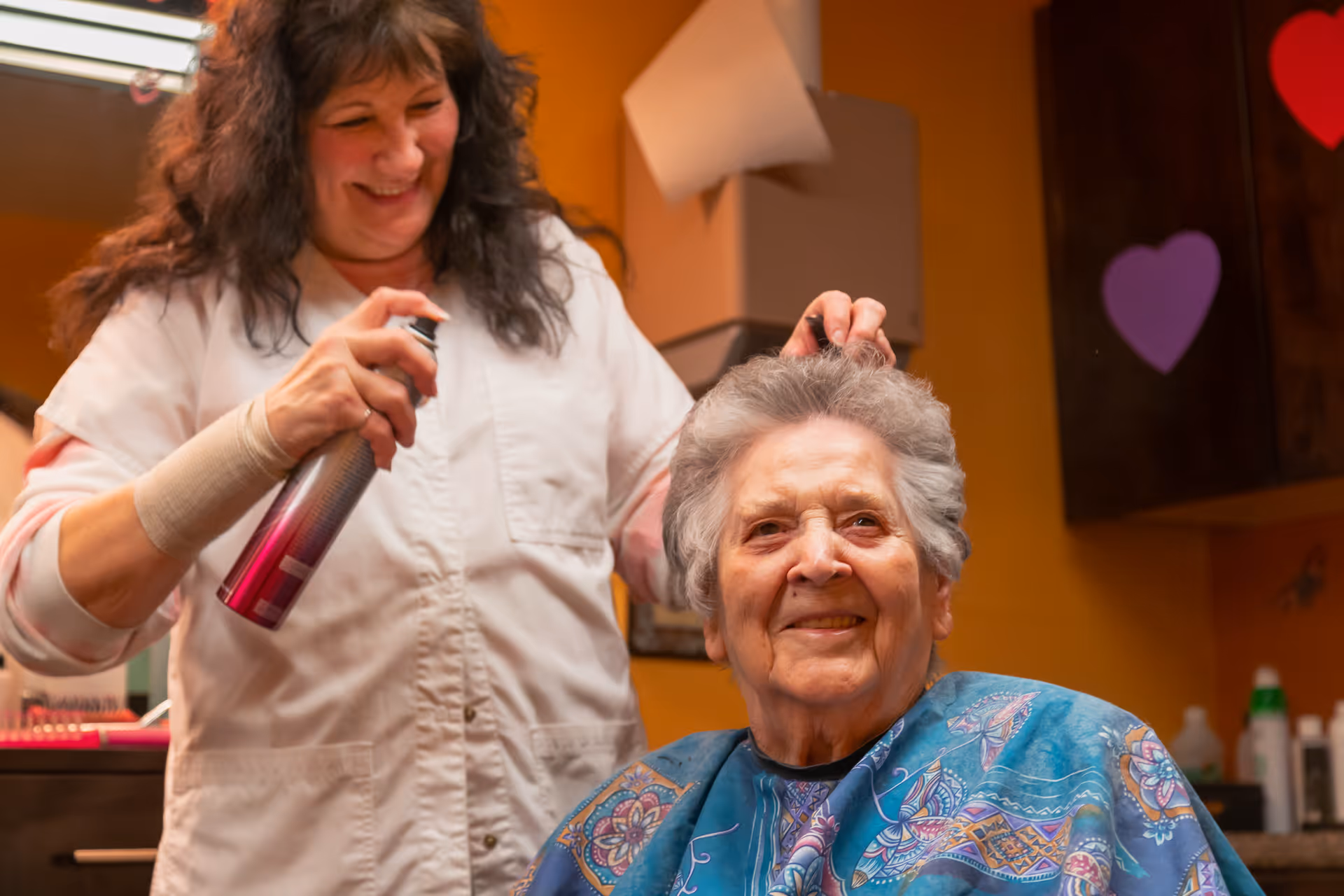 An elderly woman with gray hair is smiling while a hairstylist sprays her hair with hairspray in a warm-colored room decorated with heart shapes.