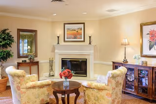 A cozy living room with two floral upholstered armchairs facing a small round wooden table with a vase of red flowers. Behind the chairs is a white fireplace with a framed landscape painting above it and two candle holders on the mantel. To the right is a wooden cabinet with glass doors, a table lamp, and a framed floral artwork on the wall. A potted plant and a mirror on a small table are visible to the left. The room has warm lighting and beige walls with a patterned carpet.