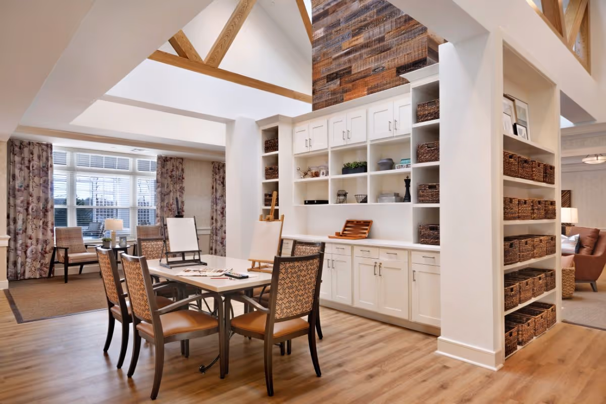 Bright communal activity room with a central table and chairs, built-in white shelving filled with wicker baskets, and large windows with patterned curtains.