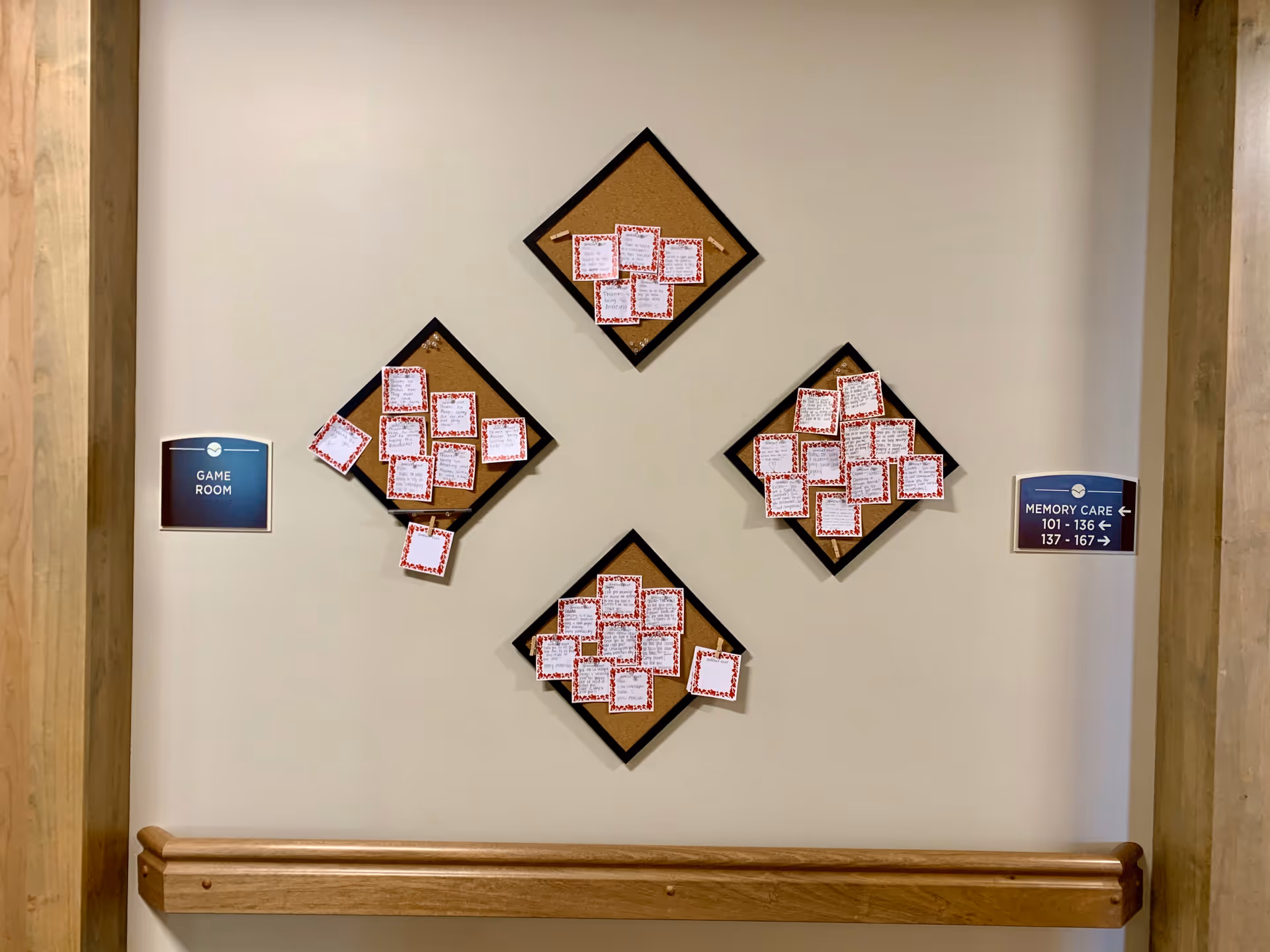 Interior hallway wall with four diamond-shaped cork boards covered in pinned notes, a wooden handrail below, and signs pointing to the Game Room and Memory Care.
