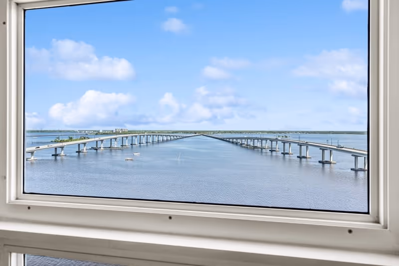 View through a window showing a long bridge extending over a body of water under a blue sky with scattered clouds.