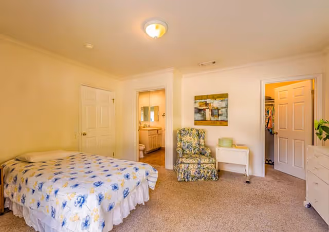 Cozy bedroom with a patterned bed, floral chair, dresser, and bathroom doorway.