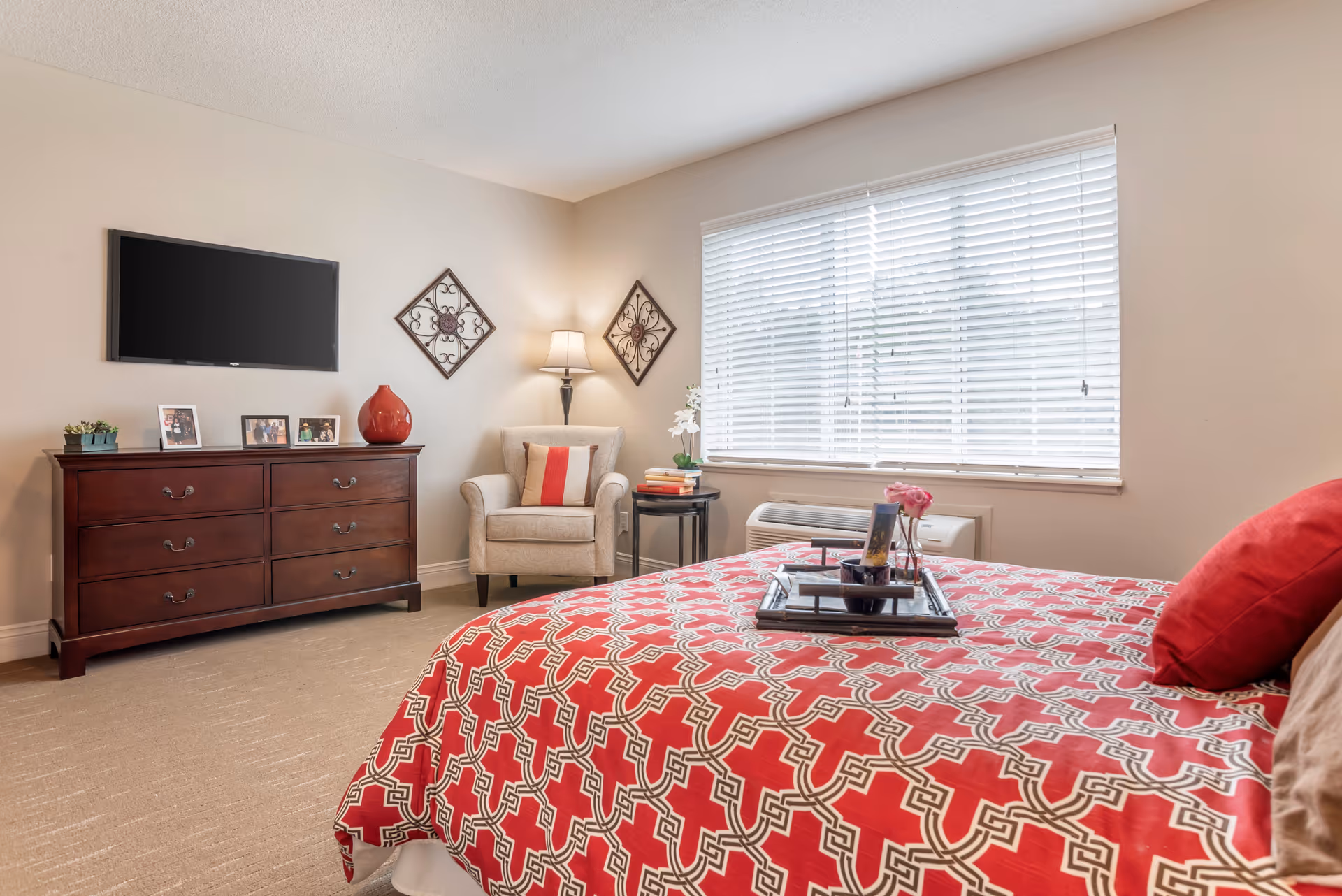 A bedroom with a large window covered by white blinds, a bed with a red and white patterned bedspread and red pillows, a wooden dresser with framed photos and a red vase, a beige armchair with a red and white pillow, a floor lamp, and two decorative wall hangings.