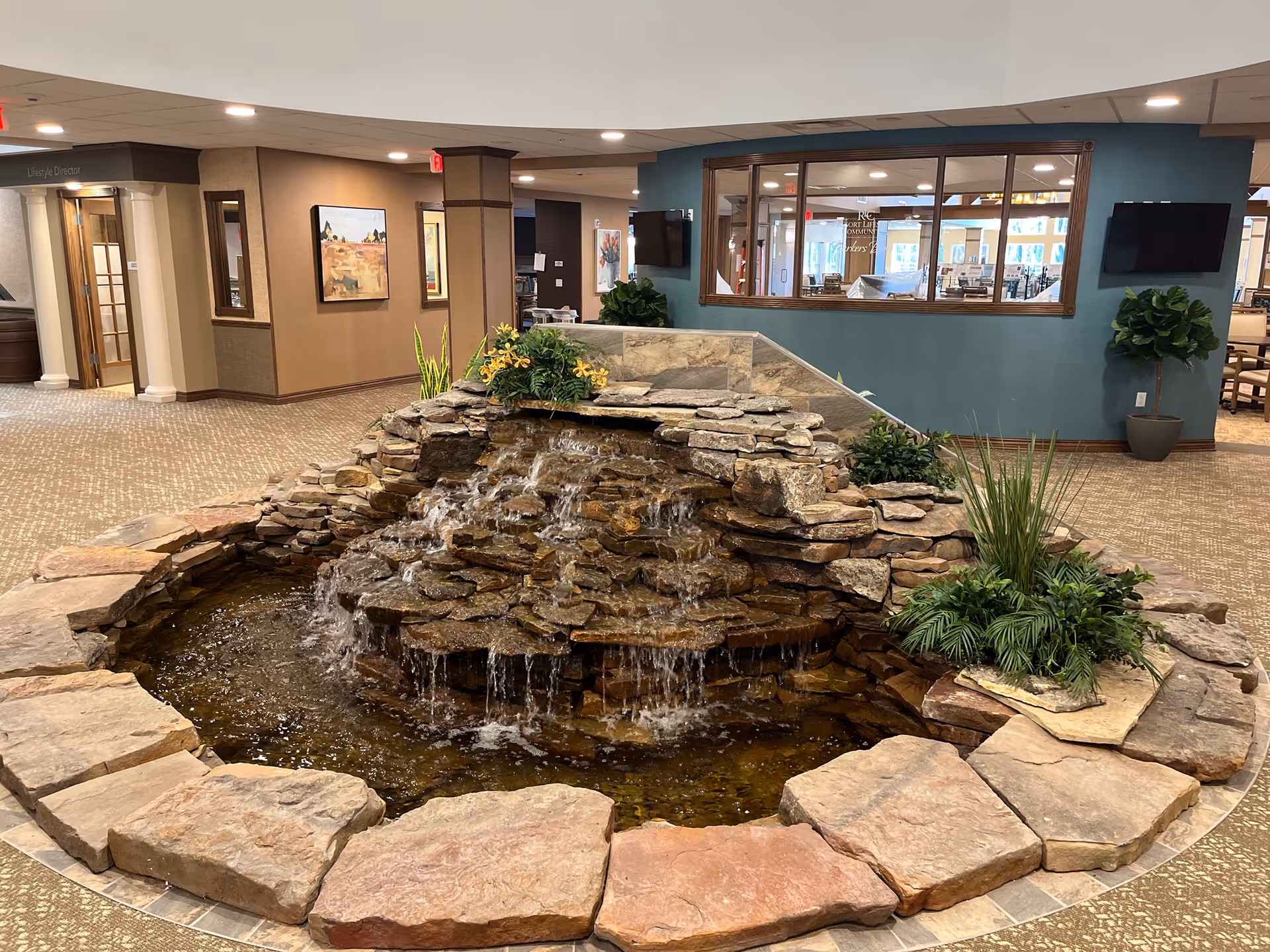 Indoor water fountain made of stacked stones with water cascading down multiple levels, surrounded by plants and large flat stones, located in a spacious room with carpeted floors, beige walls, and large windows looking into another room.