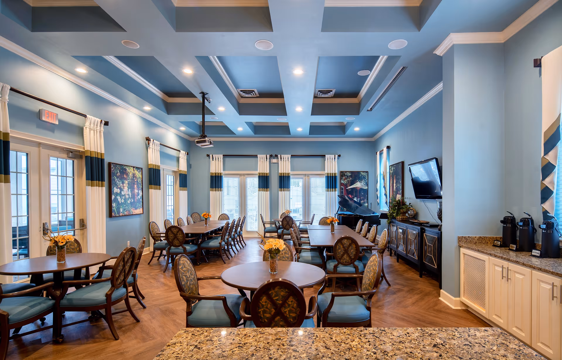 A spacious dining room with multiple round and rectangular wooden tables surrounded by cushioned chairs. The room features light blue walls, large windows with white and blue curtains, a coffered ceiling with recessed lighting, and a mounted flat-screen TV. There is a sideboard with decorative items and a coffee station with dispensers on a granite countertop.
