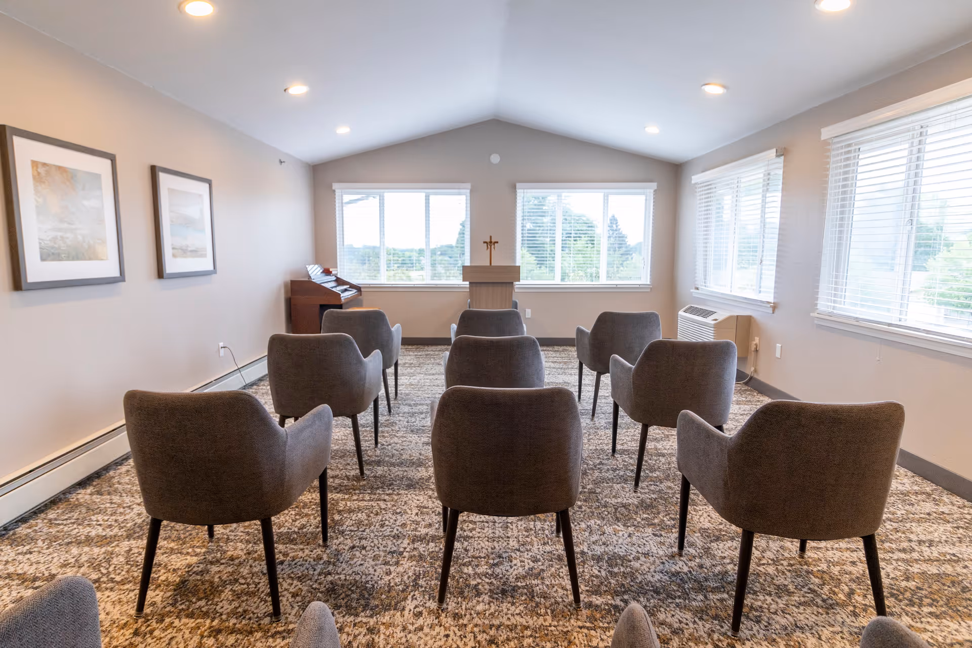 A small meeting or chapel room with two rows of gray upholstered chairs facing a wooden podium with a cross on it. The room has large windows on two walls letting in natural light, beige walls with two framed pictures, and a carpeted floor.