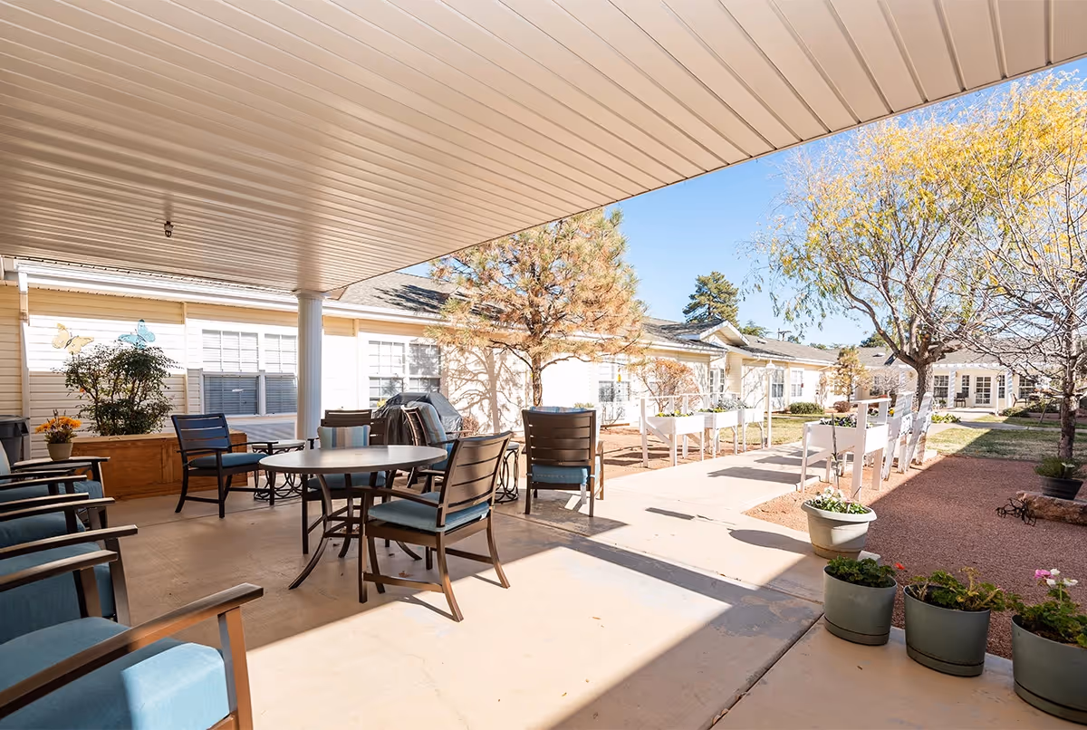 Covered outdoor patio area with several chairs and a round table, adjacent to a garden with potted plants, raised garden beds, and trees. The patio is part of a senior living facility building with light-colored siding and multiple windows.