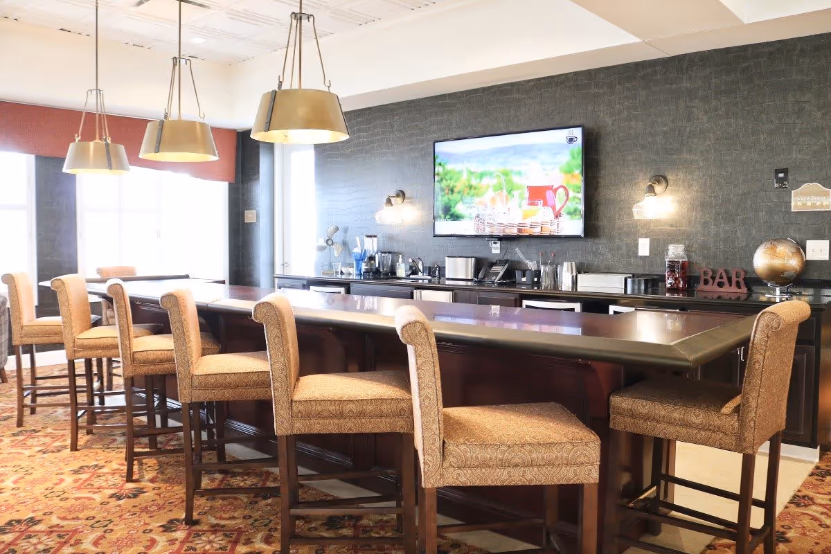 A well-lit bar area in a senior living facility with a long counter surrounded by upholstered bar stools. The back wall features a mounted flat-screen TV displaying a scenic image, various beverage appliances, and decorative items including a globe and a sign that says 'BAR'. Large windows allow natural light to fill the room.