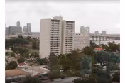 View of a tall white apartment building surrounded by trees with a city skyline and a bridge in the background under a cloudy sky.