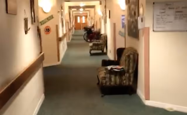 Carpeted interior hallway in a senior living facility with handrails, chairs along the walls, and a mobility scooter in the distance.