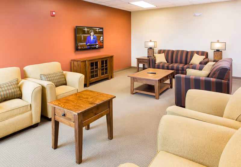 A cozy living room with beige armchairs and striped sofas arranged around wooden coffee and side tables. A flat-screen TV is mounted on an orange accent wall above a wooden cabinet. Two table lamps with beige shades are placed on side tables next to the sofas.