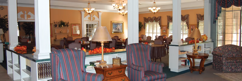 Interior view of a common area in an assisted living facility featuring striped armchairs, wooden side tables with lamps, decorative plants, and a dining area with multiple tables and chairs in the background. The room is warmly lit with chandeliers and has large windows with drapes.