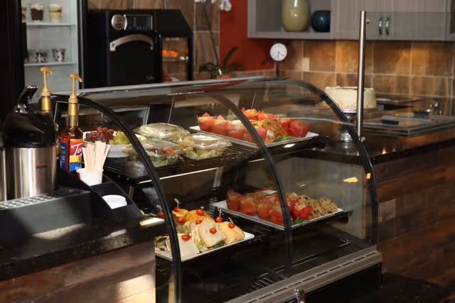 A glass display case in a kitchen or dining area containing various food items including sandwiches, salads, fruit cups, and cookies. The background shows kitchen cabinets, a countertop with a cake, and a coffee dispenser with condiments nearby.