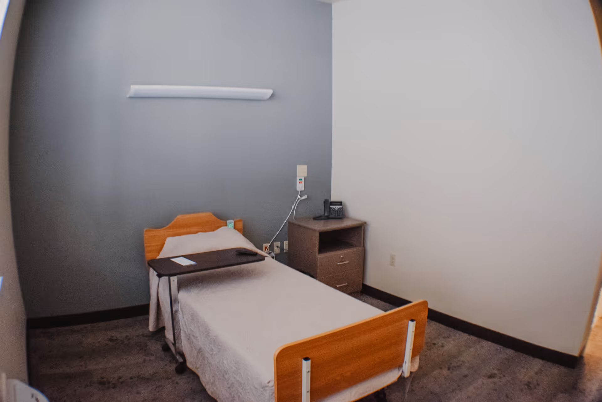 A small, simple room with a single hospital-style bed covered with a white blanket, a wooden headboard and footboard, a bedside table with a telephone, and a rolling overbed table positioned over the bed. The walls are painted in two tones, gray and white.