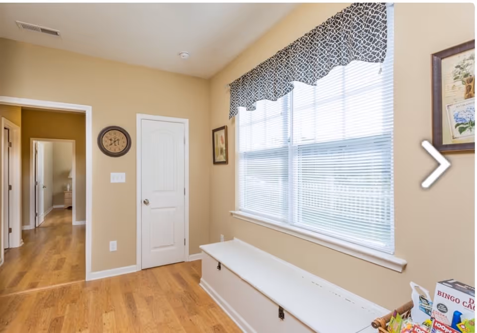A bright interior room with light beige walls and wooden flooring. There is a large window with white blinds and a patterned valance above it. Below the window is a white storage bench. On the walls are a round clock and framed artwork. A hallway with open doors is visible to the left.