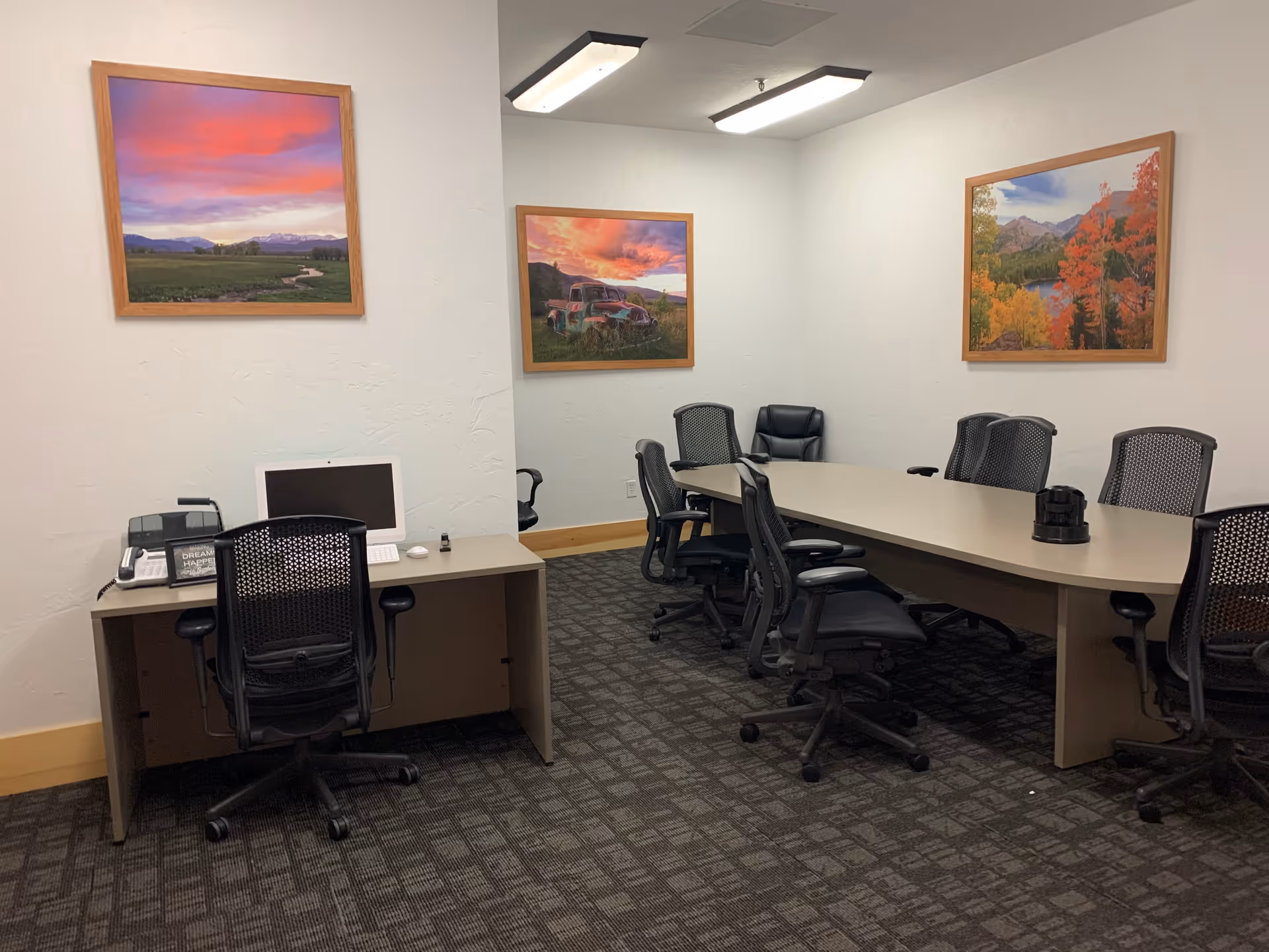 A conference room with a long table surrounded by black office chairs. There is a desk with a computer and office phone on the left side. The walls are decorated with three framed landscape photographs featuring colorful skies and nature scenes.