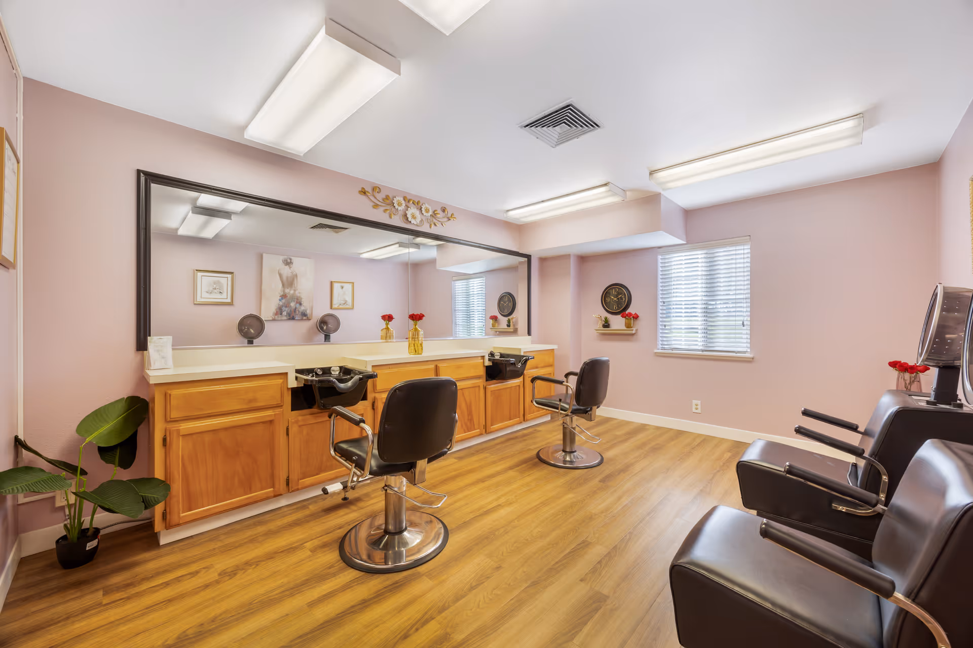 A bright and clean hair salon room with two black salon chairs in front of a large mirror mounted on a lavender wall. Below the mirror are wooden cabinets with two black hair washing sinks. The room has wood flooring, a window with blinds, and three black waiting chairs with armrests on the right side. The walls are decorated with framed artwork, a clock, and small shelves with red flowers.