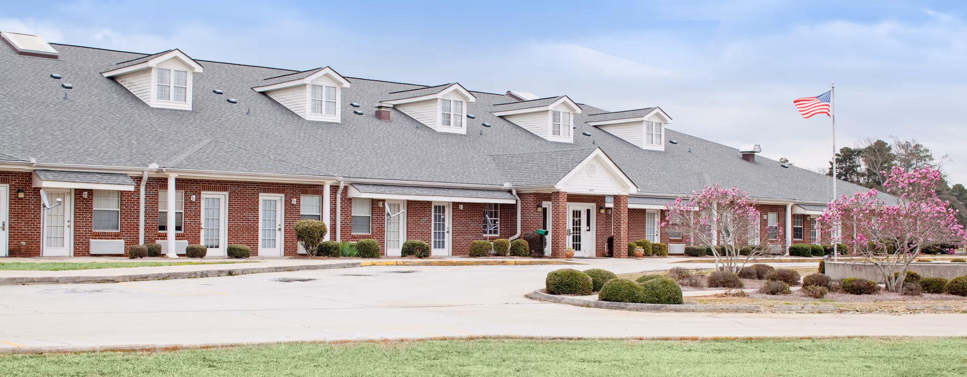 Exterior view of a single-story brick building with multiple white doors and windows, a gray shingled roof with dormer windows, an American flag on a flagpole, and landscaped bushes and flowering trees in front.