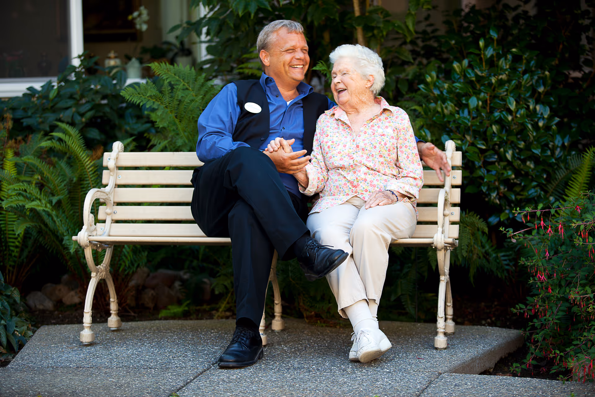 An elderly woman and a man sitting on a white bench outdoors, smiling and holding hands, surrounded by green plants and foliage.