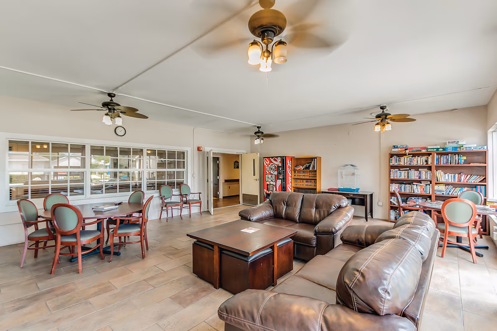 A spacious common area in Cedar Creek Assisted Living featuring two brown leather sofas arranged around a wooden coffee table. There are multiple ceiling fans with lights, a round table with green cushioned chairs, bookshelves filled with books, a vending machine, and large windows letting in natural light.
