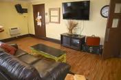 Communal living room with a leather sofa, green coffee table, wall-mounted flat-screen TV above a media cabinet, a wall clock and doors.