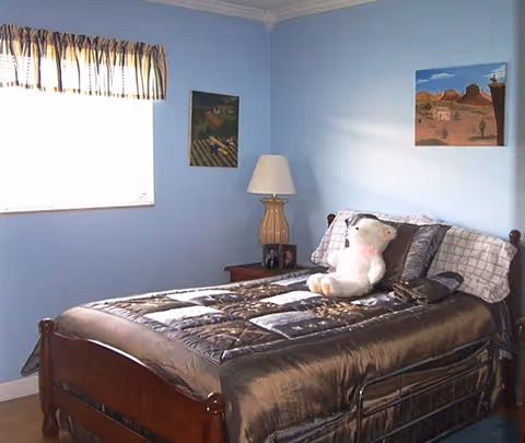 A bedroom with light blue walls featuring a wooden bed with a dark patterned comforter and pillows. A white stuffed animal is placed on the bed. There is a wooden nightstand with a lamp and framed photos next to the bed. Two paintings hang on the walls, and a window with a striped valance allows natural light into the room.