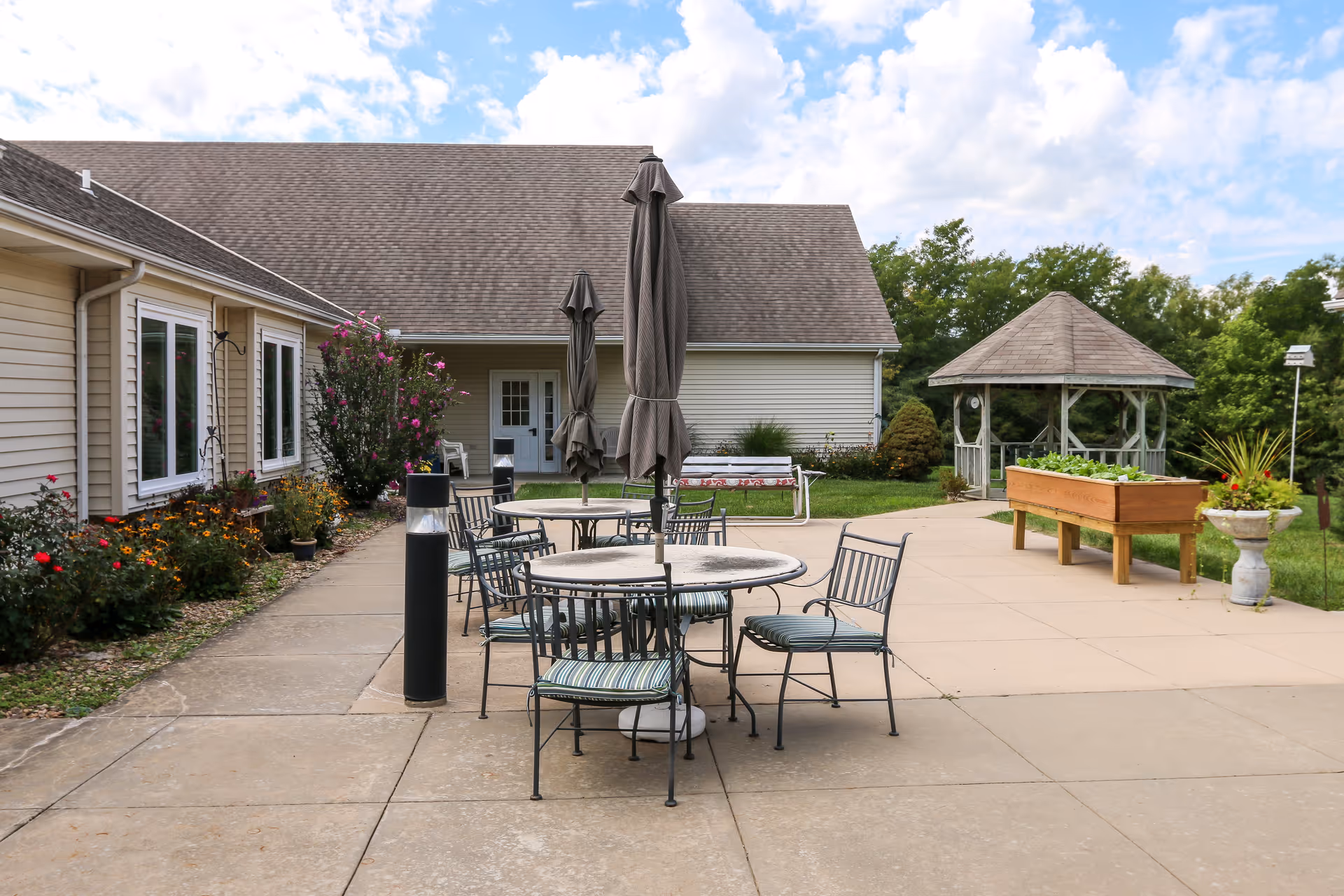 Outdoor patio with round tables and chairs under closed umbrellas, a gazebo, planter boxes, and the facility building in the background.