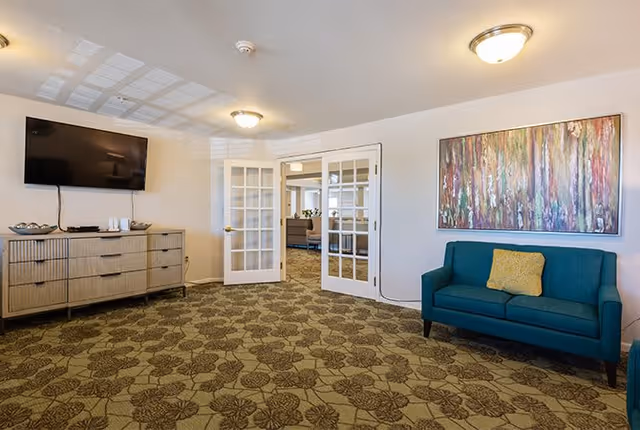Interior room with patterned carpet, a teal couch with a yellow pillow, a large abstract painting on the wall, a wooden dresser with a flat-screen TV mounted above it, and white double French doors leading to another room.