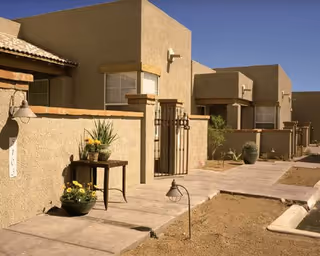 Walkway and entrance area of beige stucco apartment-style units with potted plants and a small table under a clear blue sky.