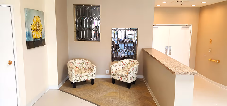 A small seating area in a hallway with two floral patterned armchairs placed on a wooden floor section. There are two decorative mirrored wall panels above the chairs and a painting of a yellow armchair on the adjacent wall. To the right is a granite countertop with a beige wall and double doors in the background.