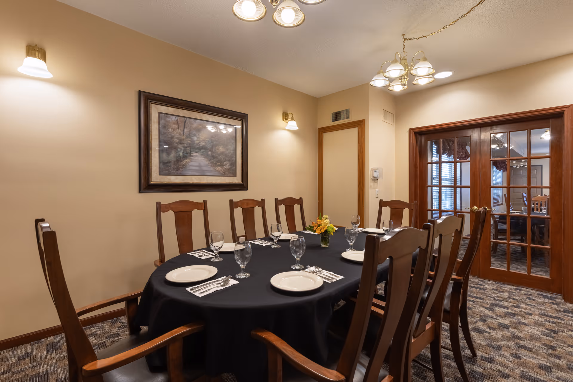 A dining room with a large oval table covered with a black tablecloth, set with white plates, silverware, and water glasses. Eight wooden chairs surround the table. The room has beige walls with a framed picture of a forest path, two wall sconces, and a chandelier overhead. There are wooden framed glass double doors leading to another room.