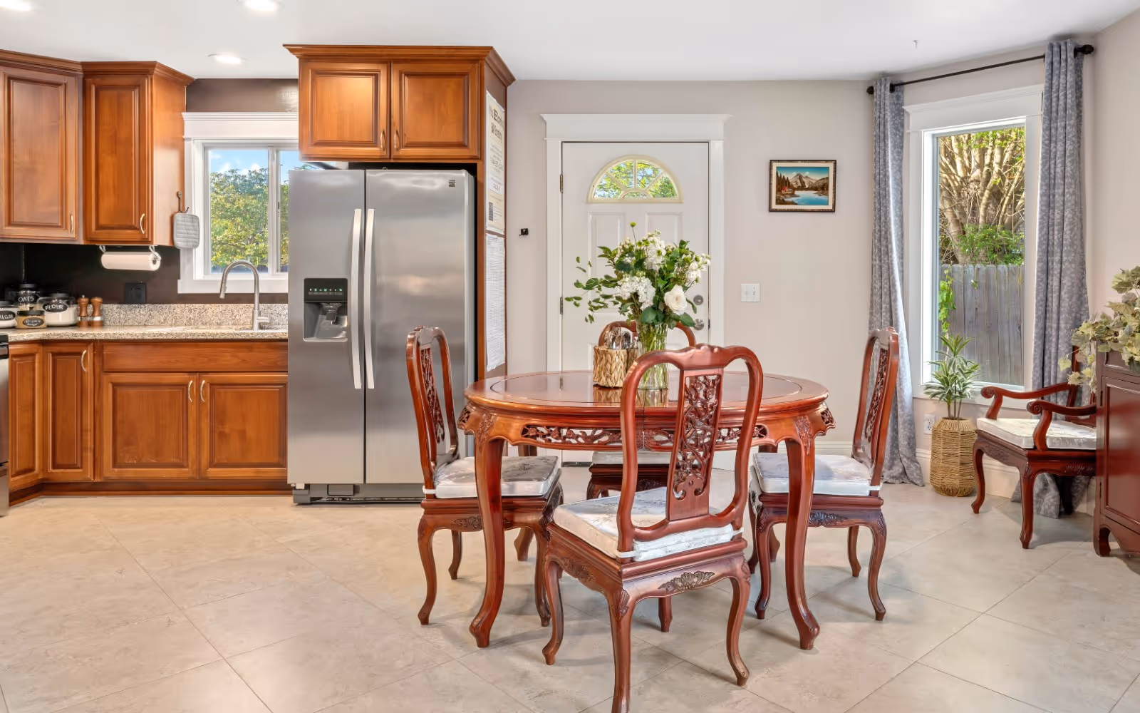 A bright kitchen and dining area featuring wooden cabinets, a stainless steel refrigerator, and a round wooden dining table with four carved wooden chairs. A vase with white flowers is placed on the table. There is a window above the sink, a door with a small window, and a large window with gray curtains letting in natural light. The floor is tiled, and there are decorative plants and a framed picture on the wall.